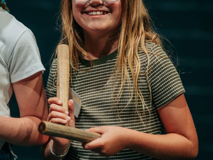 Image of A smiling girl with long blonde hair and blue glasses holds two wooden sticks, standing against a dark background. She wears a striped shirt and jeans.