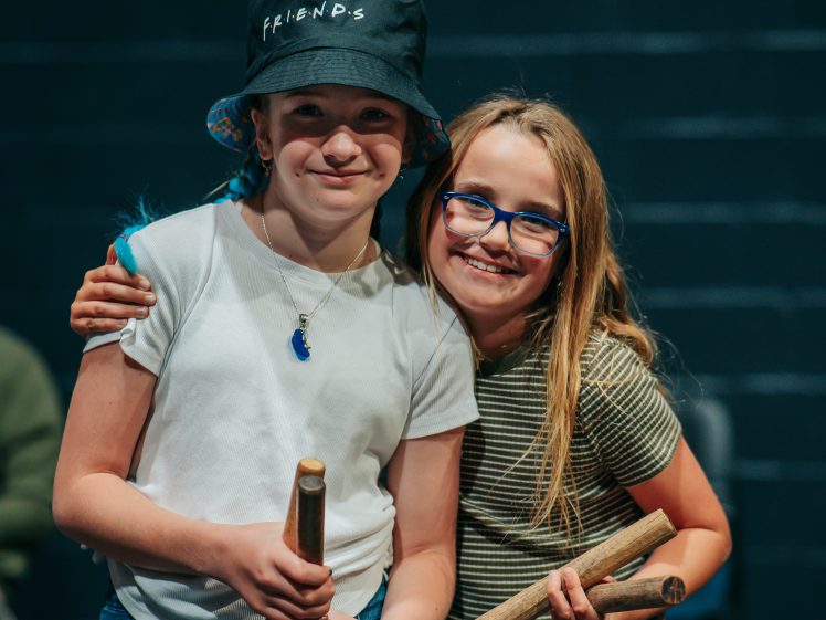 Image of Two smiling children stand close together, each holding wooden rhythm sticks. One wears a bucket hat labelled FRIENDS and a white shirt, while the other wears glasses and a striped shirt. They appear happy and friendly.