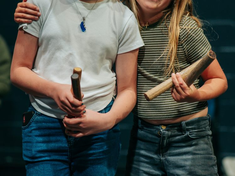 Image of Two smiling children stand close together, each holding a wooden stick. One wears a black bucket hat, white shirt, and jeans; the other has glasses, a striped shirt, and denim shorts. They appear happy and relaxed indoors.