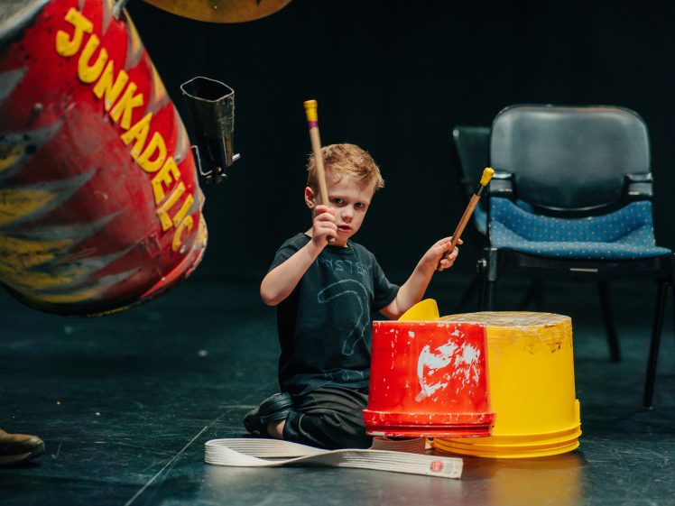 Image of A young boy kneels on the floor, playing improvised drums made from two colourful plastic buckets with drumsticks. He is focused, and a large red barrel labelled Junkadelic is visible beside him.