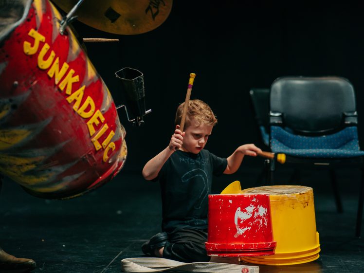 Image of A young boy sits on the floor playing drums made from colourful plastic buckets and recycled materials, with a chair and musical instruments in the background. The word JUNKAPELIC is visible on one of the drums.