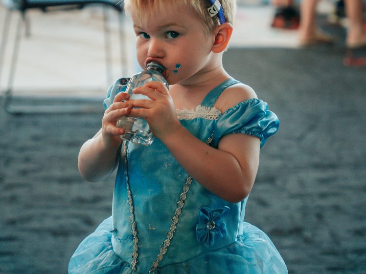 Image of A young child in a blue princess dress and purple tiara drinks from a water bottle, looking at the camera. The background is blurred with people and chairs visible.