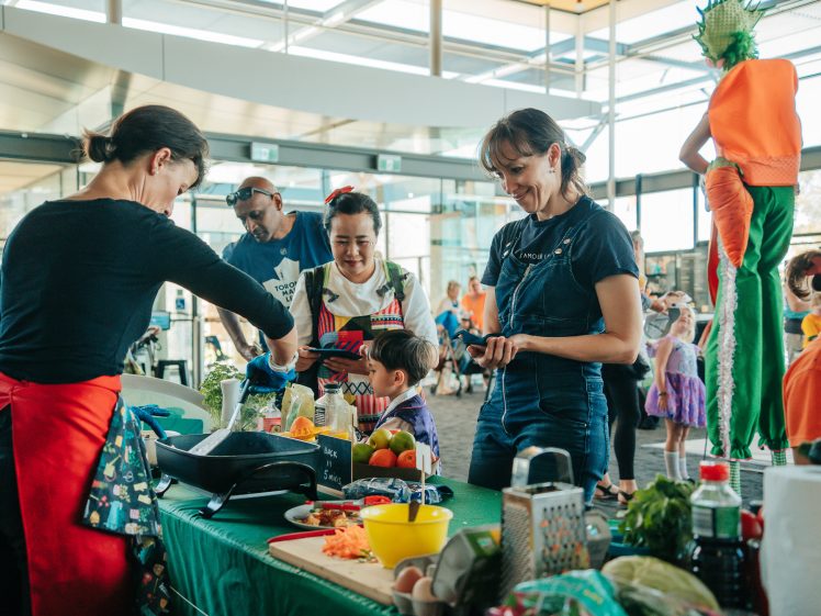 Image of A group of adults and children interact at a colourful indoor market or fair. A woman prepares food at a table with fresh produce, while others, including a child, watch and smile. A performer dressed as a carrot is seen in the background.