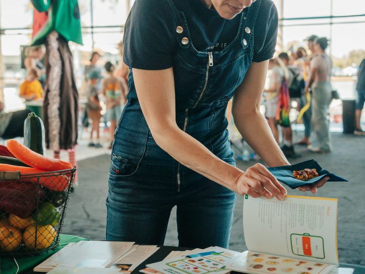 Image of A woman in dungarees stands at a table covered with leaflets and booklets, reading a small booklet. People and a person in a vegetable costume are visible in the busy background at an indoor event.
