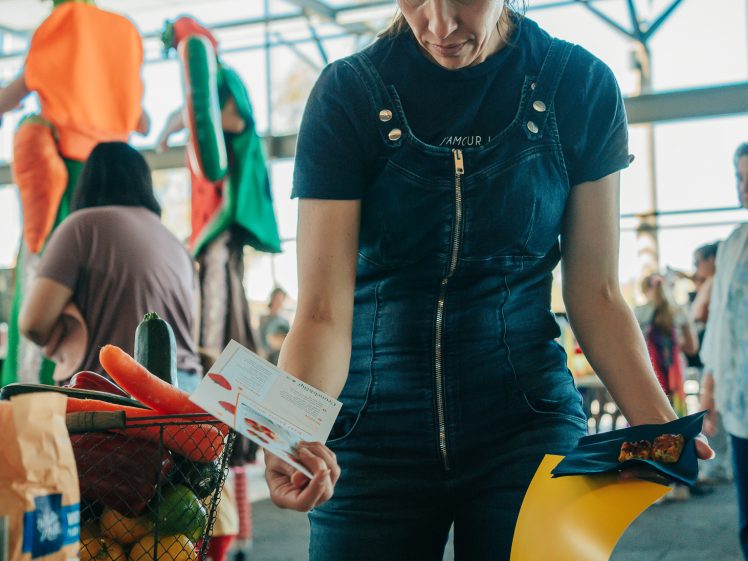 Image of A woman stands at a table reading leaflets, surrounded by informational materials and a basket of vegetables, while people in fancy dress gather in the background at a bright indoor event.