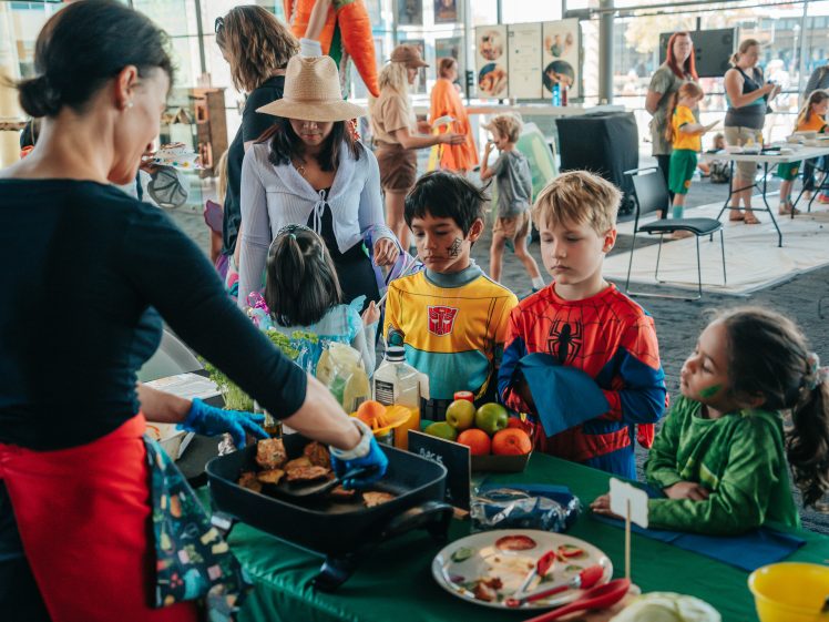 Image of A woman serves food to children dressed in colourful costumes at a festive indoor event. Kids wait in queue by a table filled with fruit and snacks, while adults and other children interact in the background.