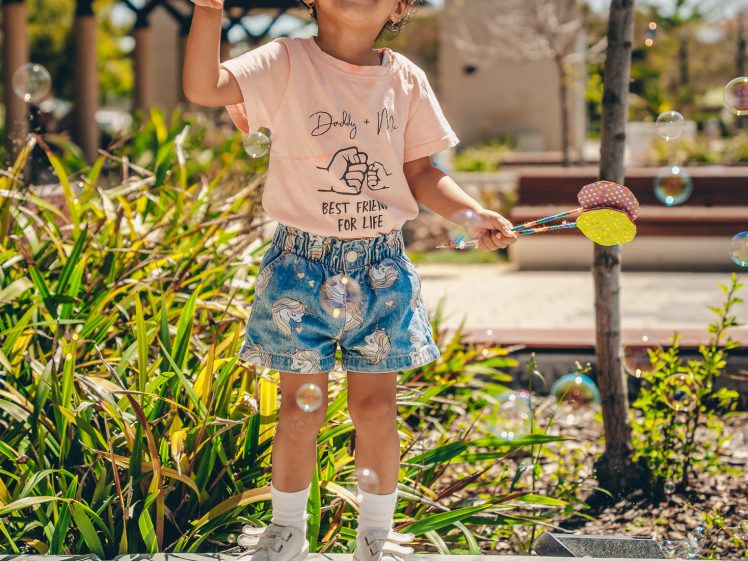 Image of A young child smiling and playing with a bubble wand outdoors, standing on a low concrete wall near green plants on a sunny day, surrounded by floating bubbles.