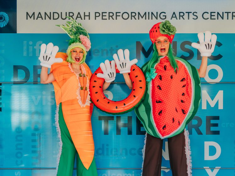 Image of Two people in colourful costumes—a carrot and a watermelon—pose with oversized white gloves in front of a blue Mandurah Performing Arts Centre backdrop. Both smile and appear enthusiastic and animated.