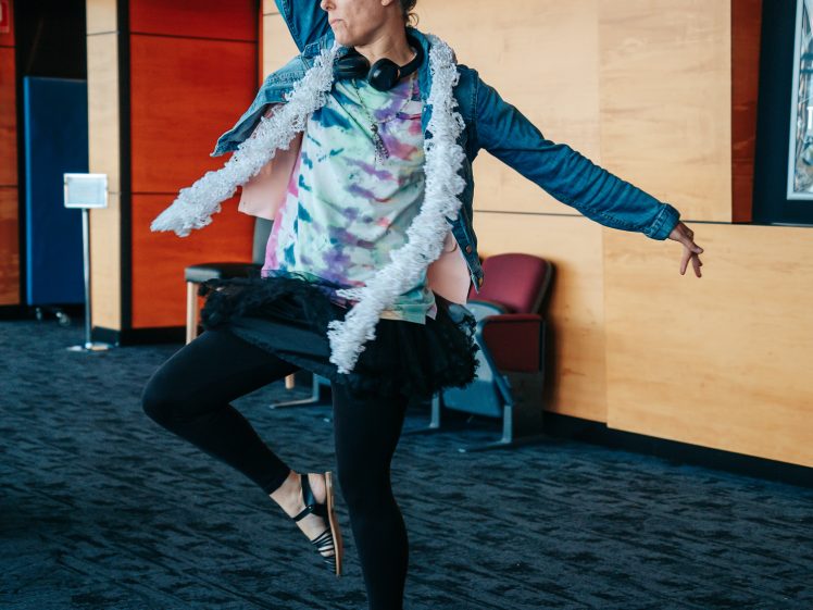 Image of A person wearing a colourful tie-dye shirt, tutu, jeans jacket, and sandals strikes a playful dance pose indoors, with one arm raised and one leg bent, in a modern room with wooden panelled walls and blue carpet.