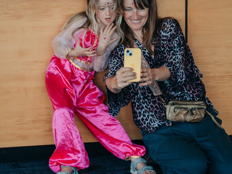 Image of A woman and a young girl in a pink costume sit against a wooden wall, smiling as they take a selfie with a yellow mobile. The girl has face paint and waves, while the woman wears sunglasses and a leopard-print top.