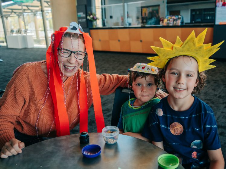 Image of A smiling adult with red ribbon headgear sits at a table with two children in colourful, homemade hats. One child wears a yellow sun hat, and the other wears a paper hat. Small containers of snacks are on the table.