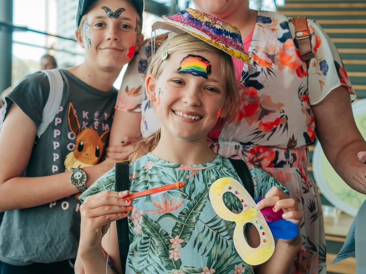 Image of A smiling woman and two children with painted faces and costumes pose indoors. One child holds a decorated mask, and the other wears an Eevee shirt. The group looks cheerful and festive.