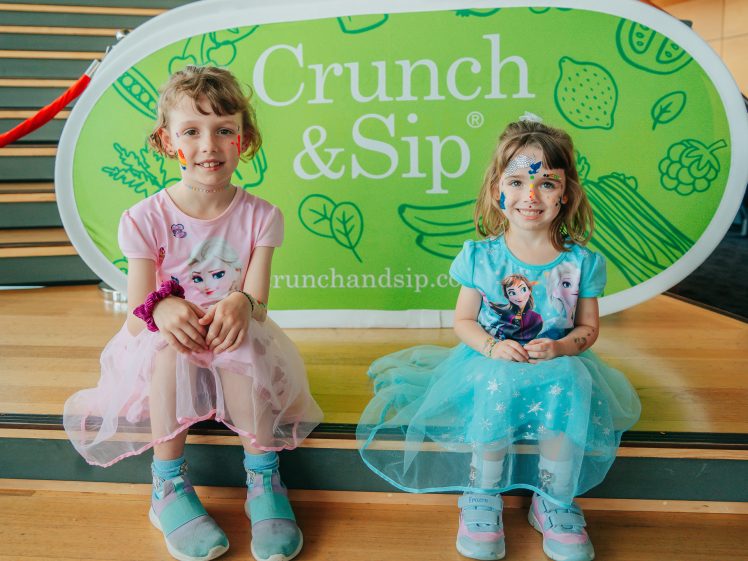 Image of Two young girls in pastel-coloured Frozen-themed dresses sit on wooden steps, smiling, with face paint. Behind them is a green Crunch & Sip sign with fruit and vegetable illustrations.