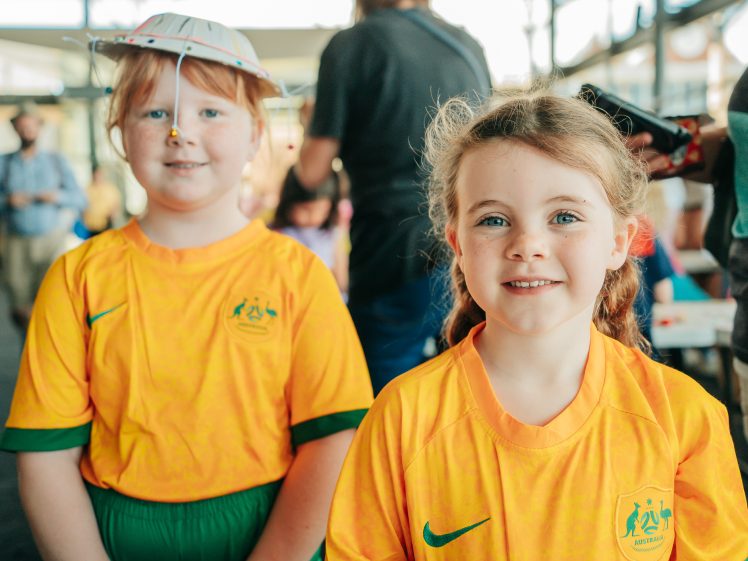 Image of Two young girls wearing yellow Australia football shirts stand indoors. One has a homemade hat and face paint; both smile at the camera. People and tables are visible in the blurred background.