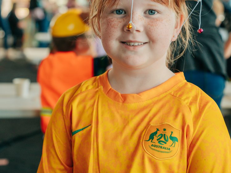 Image of A smiling child with red hair and freckles wears a yellow Socceroos shirt and a homemade hat decorated with beads and strings. The background is blurred, showing other people and activity indoors.