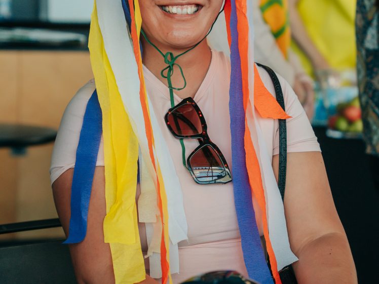 Image of A smiling person wearing a colourful hat with long ribbon streamers and a mask on top sits indoors. They have sunglasses hanging from their shirt and a decorative blue glove on the table in front of them.