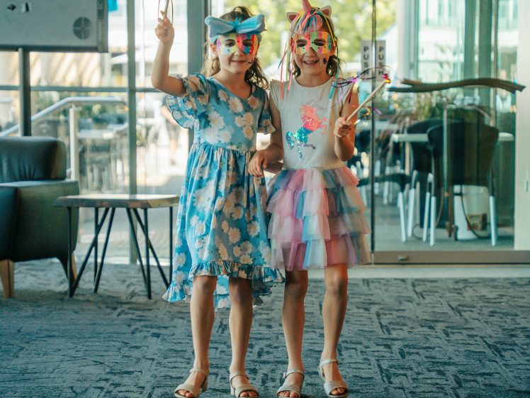 Image of Two young girls stand indoors wearing colourful party masks and holding wands. One wears a blue floral dress and the other a rainbow skirt with a unicorn headband. They smile and pose together in a bright, modern room.