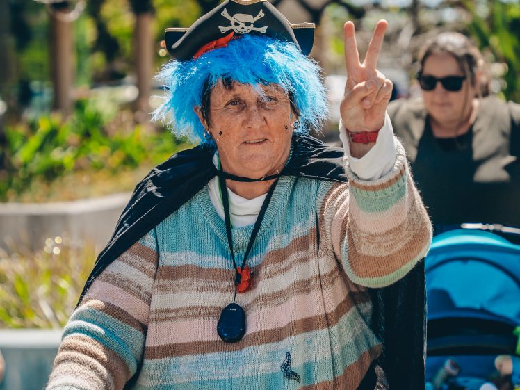 Image of An older woman wearing a blue wig, pirate hat, and striped jumper makes a peace sign while using a Zimmer frame. She is outdoors, with another woman and a pushchair visible in the background.