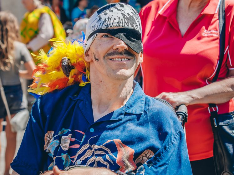 Image of A smiling man in a wheelchair, dressed as a pirate with an eye patch and feathered hat, is pushed by a cheerful woman in a red shirt at a lively outdoor event.
