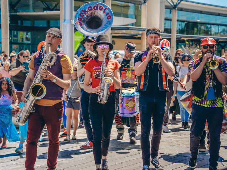 Image of A lively street band with four musicians playing brass instruments marches outdoors, followed by a crowd. The musicians wear colourful, eclectic clothing and sunglasses, with people watching and enjoying the performance.