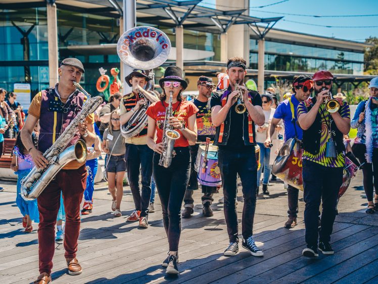 Image of A lively street band marches outdoors, playing brass and woodwind instruments. They are followed by people, some in colourful clothing, enjoying the parade on a sunny day near modern buildings.