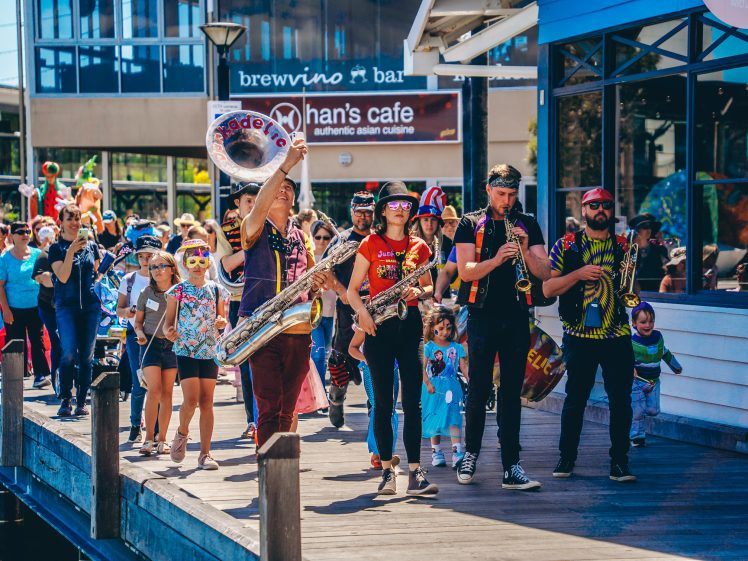Image of A lively street band with brass instruments leads a colourful parade of adults and children along a promenade, passing cafés and onlookers under sunny, festive weather.