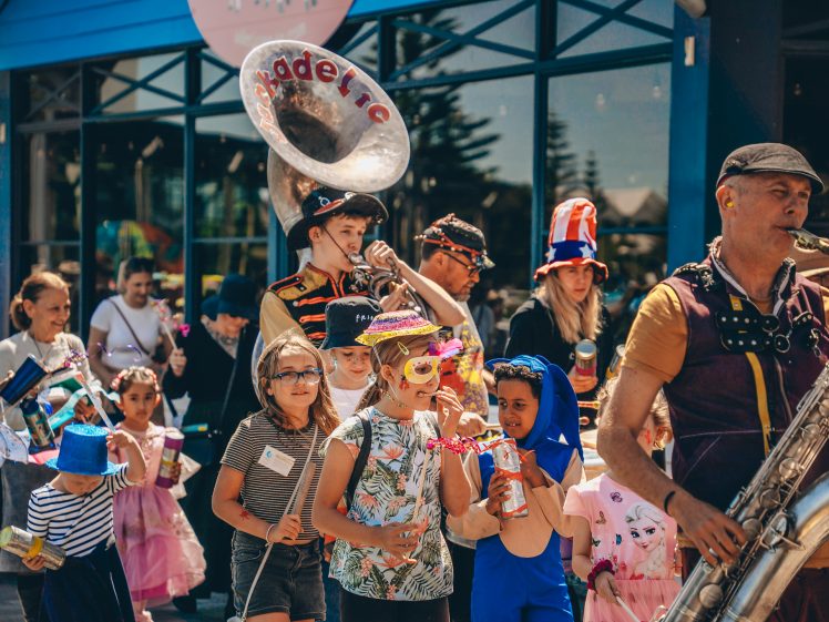 Image of A lively parade featuring musicians playing brass instruments and children in colourful costumes and masks marching together outside a building on a sunny day.