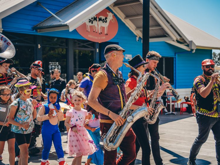 Image of A lively street band with colourful costumes plays music as children in fancy dress parade alongside them outside a bright blue building on a sunny day.