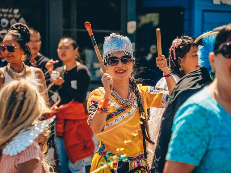 Image of A smiling girl in a bright yellow traditional outfit and silver headdress plays sticks among a crowd during a lively outdoor celebration. Other people in colourful clothing surround her, enjoying the festivities.
