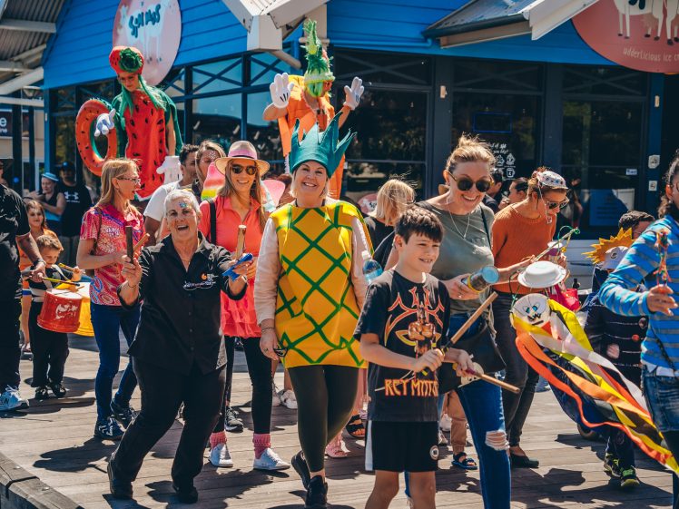 Image of A lively group of people, some in colourful costumes like a pineapple and an octopus, parade outdoors on a sunny day, smiling and waving flags. Children and adults enjoy the festive atmosphere near a café.