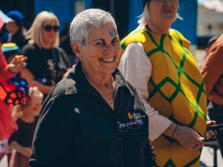 Image of An older woman with short grey hair and blue face paint smiles at an outdoor event. She wears a black shirt, and people in colourful costumes, including one in a pineapple suit, are in the background.