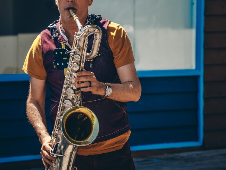 Image of A man wearing a cap and casual clothes plays a saxophone outdoors in front of a building with blue window frames and wooden cladding.