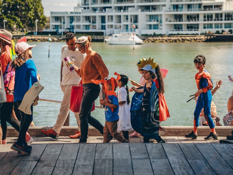 Image of A group of children in colourful costumes, including superheroes and fairies, walk in a parade along a waterfront promenade, accompanied by adults on a sunny day. Modern buildings and a boat are visible in the background.