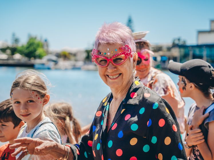 Image of An older woman with pink hair and bright pink, heart-shaped glasses smiles at a waterfront event, wearing a colourful polka-dot shirt, surrounded by children and adults with face paint.