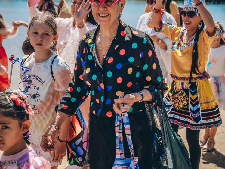 Image of A smiling elderly woman in a colourful polka-dot shirt and pink sunglasses leads a joyful outdoor parade with people in festive costumes, including children in dresses, near a waterfront.