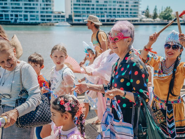 Image of A lively group of people, including children and adults in colourful costumes, walk outdoors near water and high-rise buildings, celebrating with smiles, fans, and musical instruments.