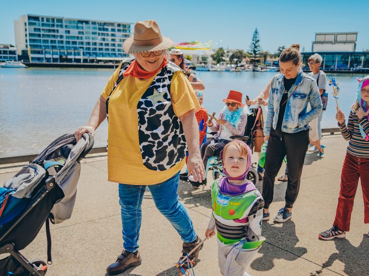 Image of A group of people, including children and adults, walk by a waterfront. One adult wears a cow-print waistcoat and hat, holding hands with a child dressed as Buzz Lightyear. Others wear colourful costumes and push a pram on a sunny day.