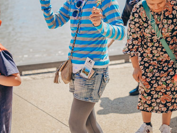 Image of A woman in a striped blue hoodie, denim shorts, and grey tights smiles while holding a tin and a decorated stick. She has fake white facial hair and spectacles, standing outdoors near water with others around her.