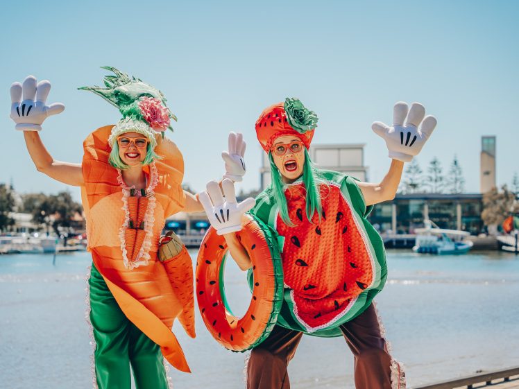 Image of Two people dressed in whimsical carrot and watermelon costumes, wearing large cartoon gloves and glasses, happily posing by a waterfront on a sunny day with blue sky and buildings in the background.