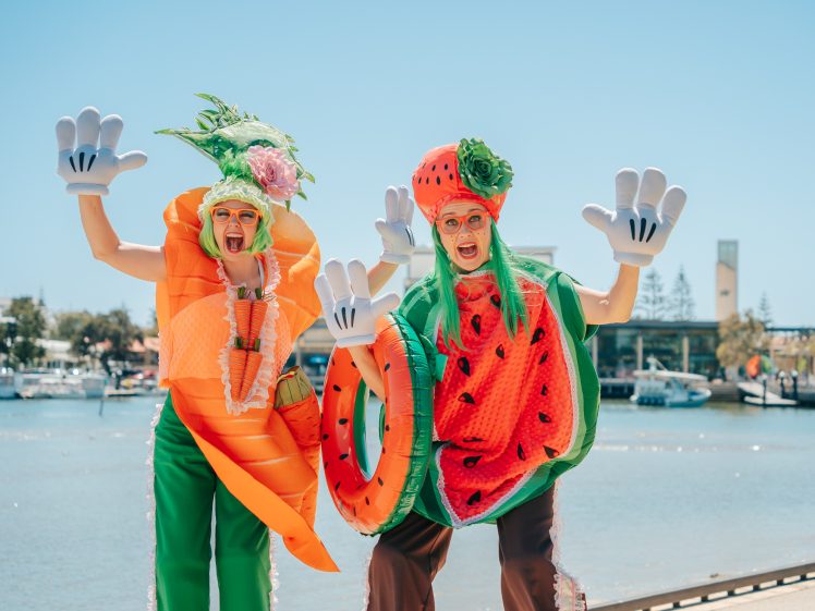 Image of Two people in funny fruit and vegetable costumes—one as a carrot, the other as a watermelon—pose playfully with big cartoon gloves near a waterfront on a sunny day.