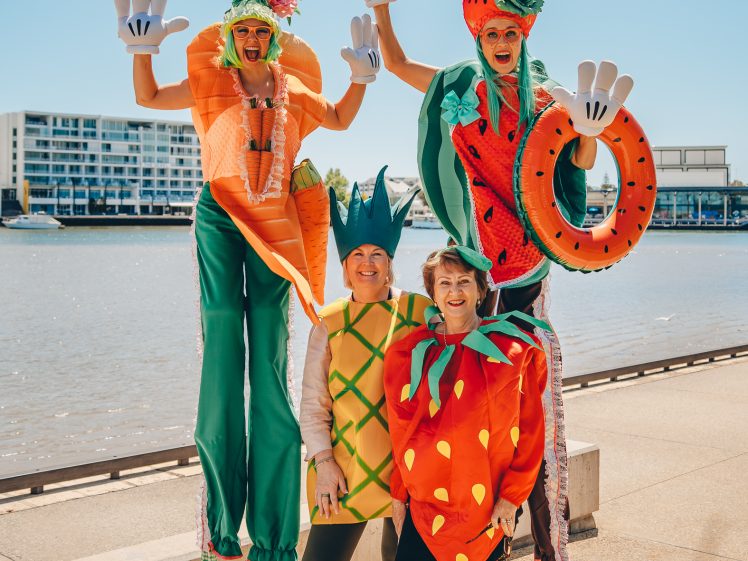 Image of Four people dressed in colourful fruit and vegetable costumes—carrot, pineapple, strawberry, and watermelon—posing happily by a riverside on a sunny day, with two on stilts and two standing on the ground.