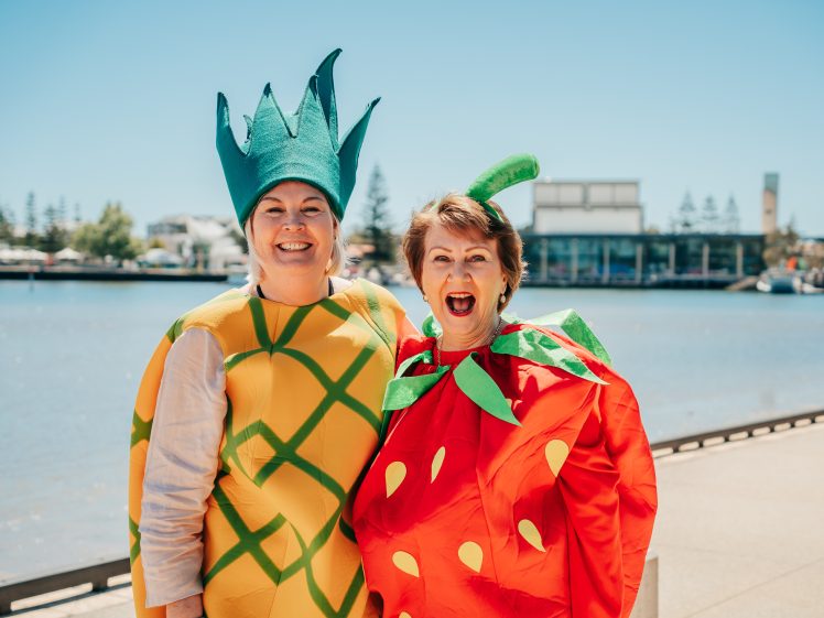 Image of Two people stand by a waterfront on a sunny day, smiling and wearing bright costumes—one dressed as a pineapple and the other as a strawberry. Buildings and trees are visible in the background.