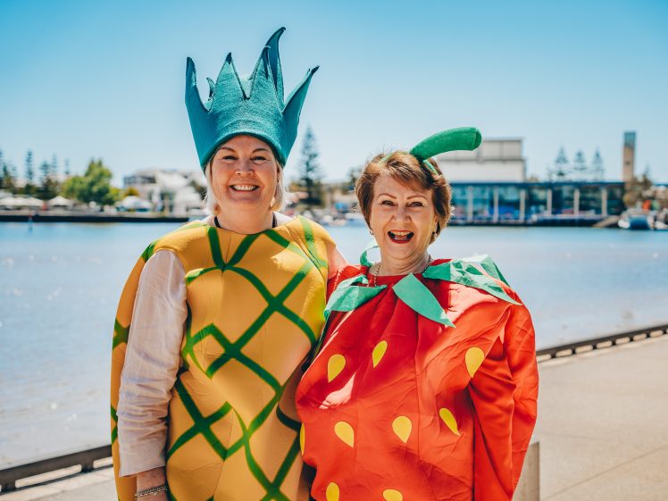 Image of Two women smiling and posing outdoors by the water, dressed in bright fruit costumes—one as a pineapple and the other as a strawberry—on a sunny day with buildings and trees in the background.