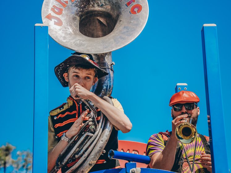 Image of Two musicians perform outdoors on a blue structure. One plays a sousaphone labelled Junkadelic; the other plays a trumpet. Both wear colourful clothing and hats, with a clear blue sky in the background.