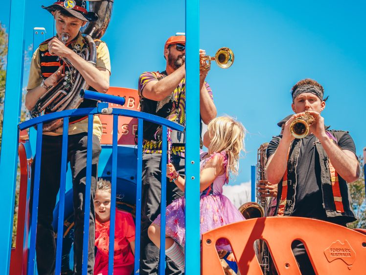 Image of A band plays brass instruments on a colourful playground structure, whilst two children in bright clothing watch and participate, against a clear blue sky.