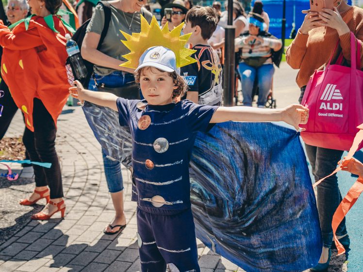 Image of A child dressed as the solar system, with a sun headpiece and planet-themed outfit, stands with arms outstretched at an outdoor event surrounded by people.