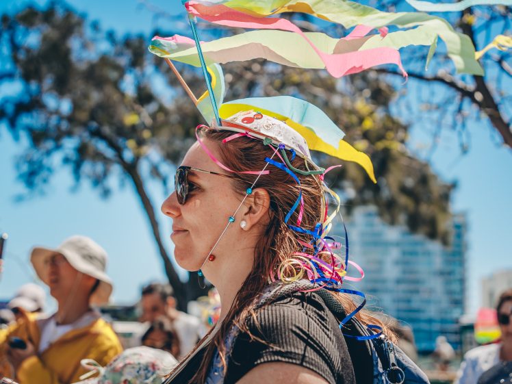 Image of A woman wearing sunglasses and a colourful headpiece with ribbons and streamers stands outdoors at a sunny event. People and trees are visible in the background under a clear blue sky.
