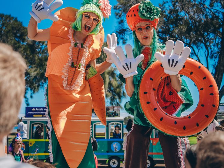 Image of Two performers on stilts dressed in colourful vegetable and fruit costumes, including a carrot and a watermelon, wave oversized gloved hands at a group of children outdoors on a sunny day.