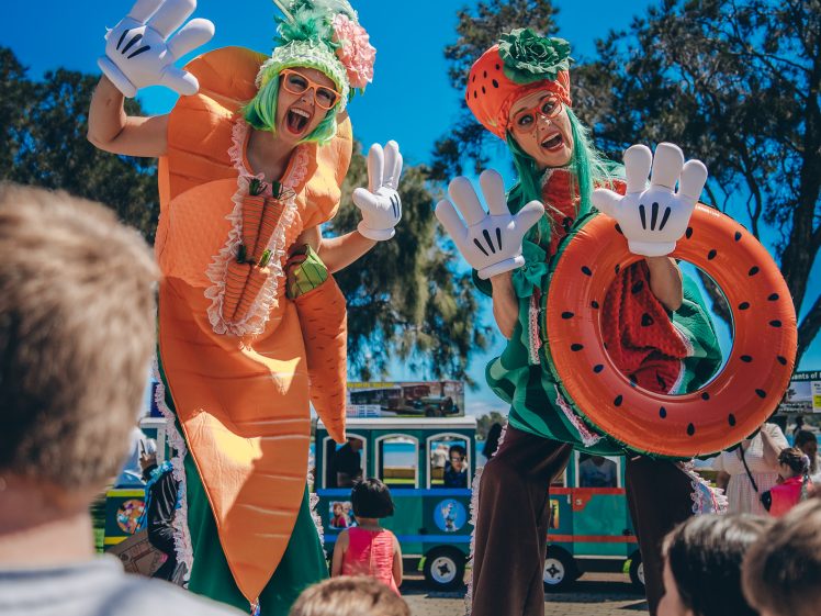 Image of Two performers on stilts dressed in colourful fruit and vegetable costumes wave playfully at a crowd of children at an outdoor event, with a small train and trees visible in the background under a clear blue sky.