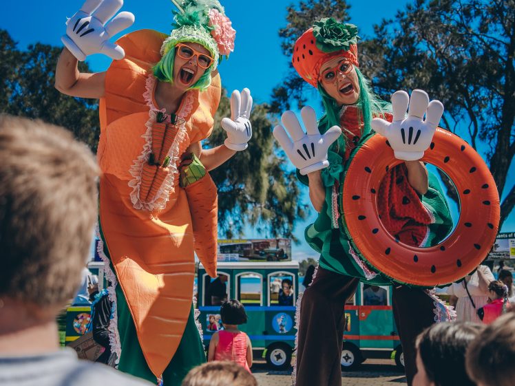 Image of Two performers on stilts dressed in whimsical fruit and vegetable costumes entertain a group of children outdoors on a sunny day, with a small train and trees in the background.